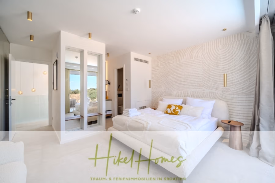 Schlüsselfertige Meerblick Villa SEA in Sutivan (Insel Brac) – 3 SZ + Bad, Pool, Sommerküche, … - Ein modernes Schlafzimmer mit großem Bett, strukturierten weißen Wänden und goldenen Leuchten. Es gibt einen Spiegelschrank, einen hölzernen Beistelltisch und gefaltete Handtücher auf dem Bett. Natürliches Licht fällt durch ein Fenster mit transparenten Vorhängen in den Raum. (42991)
