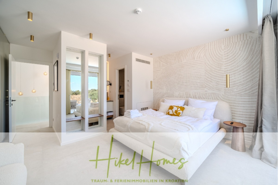 Schlüsselfertige Meerblick Villa SEA in Sutivan (Insel Brac) – 3 SZ + Bad, Pool, Sommerküche, … - Helles, modernes Schlafzimmer mit einem großen weißen Bett, goldenen Akzentkissen, strukturierter Wandkunst, Spiegelschrank und raumhohen Fenstern mit Aussicht. Der Schriftzug "Hikel Homes" und der deutsche Text erscheinen am unteren Rand. (45016)