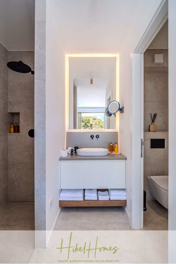 Schlüsselfertige Meerblick Villa SEA in Sutivan (Insel Brac) – 3 SZ + Bad, Pool, Sommerküche, … - Modernes Badezimmer mit einem hinterleuchteten Spiegel über einem schwebenden Waschtisch mit Handtüchern und Toilettenartikeln. Links befindet sich eine verglaste Dusche mit schwarzem Duschkopf. Die Toilette ist rechts durch eine offene Tür teilweise sichtbar. (43002)