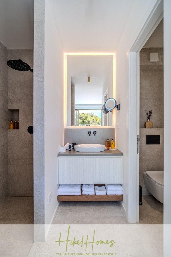 Schlüsselfertige Meerblick Villa SEA in Sutivan (Insel Brac) – 3 SZ + Bad, Pool, Sommerküche, … - Modernes Badezimmer mit einem großen beleuchteten Spiegel über dem Waschbecken, minimalistischen Armaturen, offenen Regalen mit gefalteten Handtüchern darunter und einem Duschbereich mit schwarzen Armaturen auf der linken Seite. Eine Toilette ist durch eine Tür auf der rechten Seite sichtbar. (45028)