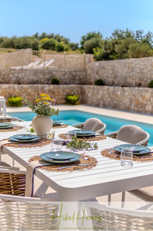 Schlüsselfertige Meerblick Villa SEA in Sutivan (Insel Brac) – 3 SZ + Bad, Pool, Sommerküche, … - Ein sonnenbeschienener Esstisch im Freien, gedeckt mit Tellern, Gläsern und einer Blumenvase neben einem klaren Swimmingpool, mit Steinmauern und Grünpflanzen im Hintergrund. (45019)