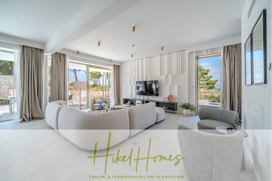 Schlüsselfertige Meerblick Villa SEA in Sutivan (Insel Brac) – 3 SZ + Bad, Pool, Sommerküche, … - Ein modernes, helles Wohnzimmer mit geschwungenen Sofas, einem Fernseher an einer strukturierten Wand, großen Fenstern und Türen zu einem Innenhof mit Meerblick. Der Text "Hikel Homes: Traum- & Ferienimmobilien in Kroatien" ist am unteren Rand eingeblendet. (45023)