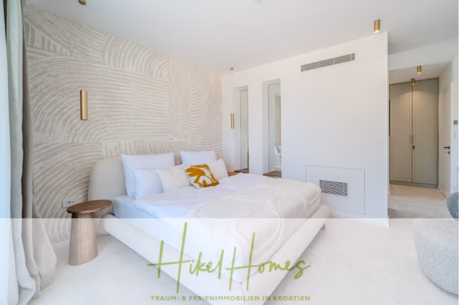 Schlüsselfertige Meerblick Villa SEA in Sutivan (Insel Brac) – 3 SZ + Bad, Pool, Sommerküche, … - Modernes Schlafzimmer mit einem weichen weißen Bett, einer gemusterten Akzentwand und minimalistischen goldenen Leuchten. Ein kleiner Beistelltisch aus Holz steht neben dem Bett. Das sanfte, neutrale Dekor schafft eine ruhige Atmosphäre. Unten ist der Schriftzug „Hikel Homes“ zu sehen. (43001)