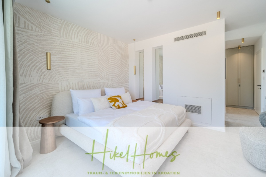 Schlüsselfertige Meerblick Villa SEA in Sutivan (Insel Brac) – 3 SZ + Bad, Pool, Sommerküche, … - Helles, modernes Schlafzimmer mit weißem Polsterbett, gelbem Akzentkissen, strukturierter Tapete, minimalem Dekor, goldenen Leuchten und natürlichem Licht aus großen Fenstern. Der Schriftzug "Hikel Homes" ist am unteren Rand eingeblendet. (45027)