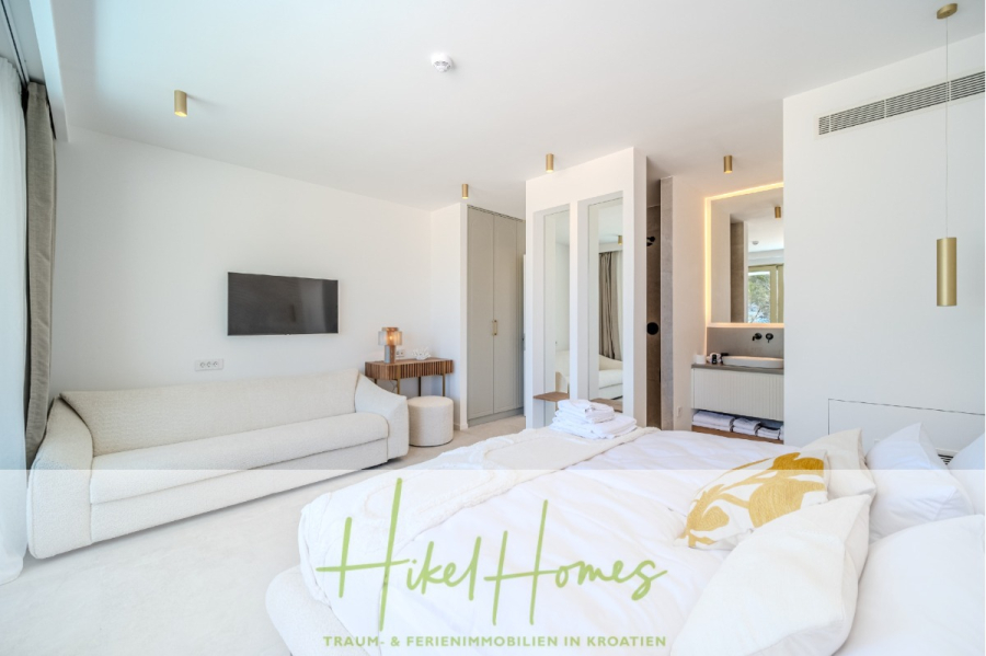 Schlüsselfertige Meerblick Villa SEA in Sutivan (Insel Brac) – 3 SZ + Bad, Pool, Sommerküche, … - Helles, modernes Schlafzimmer mit großem, ungemachtem Bett, weißem Sofa, an der Wand befestigtem Fernseher und offenem Badbereich. Goldfarbene Akzente und minimalistisches Dekor sorgen für ein sauberes, luftiges Gefühl. Der Schriftzug "Hikel Homes" ist am unteren Rand sichtbar. (45021)