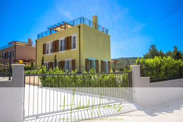 Ein modernes, gelbes, zweistöckiges Haus mit braunen Fensterläden, einer Dachterrasse und einer Auffahrt mit Tor, umgeben von Grün unter einem klaren, blauen Himmel. - Villa im Mittelmeerstil mit 3 SZ, Pool, Dachterrasse mit Meerblick (2)