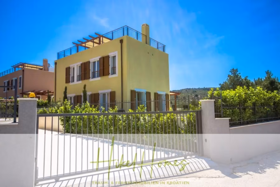 Villa im Mittelmeerstil mit 3 SZ, Pool, Dachterrasse mit Meerblick (2) - Ein zweistöckiges gelbes Haus mit Holzfensterläden und Dachterrasse liegt mitten im Grünen unter einem strahlend blauen Himmel. Ein Metalltor umschließt das Grundstück. Im Hintergrund ist ein weiteres, ähnliches Haus zu sehen. (41871)