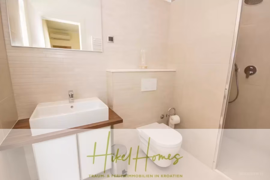 Villa im Mittelmeerstil mit 3 SZ, Pool, Dachterrasse mit Meerblick (2) - Ein modernes Badezimmer mit Waschbecken auf einer Holzplatte, Toilette und ebenerdiger Dusche. Die Fliesen sind hell, über der Toilette befindet sich ein kleines Regal. Im unteren Bereich ist das Logo von Hikel Homes zu sehen. (41886)