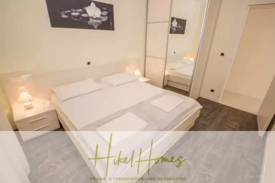 Villa im Mittelmeerstil mit 3 SZ, Pool, Dachterrasse mit Meerblick (2) - Ein modernes Schlafzimmer verfügt über ein ordentlich gemachtes Bett mit weißer Bettwäsche und zwei Kissen. Es gibt Nachttische mit Lampen auf beiden Seiten, einen großen Spiegelschrank und ein florales Kunstwerk über dem Bett. Unten steht der Text „Hikel Homes“. (41885)