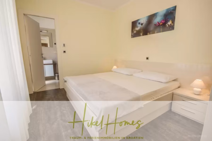 Villa im Mittelmeerstil mit 3 SZ, Pool, Dachterrasse mit Meerblick (2) - Ein minimalistisches Schlafzimmer mit Doppelbett, weißer Bettwäsche und Nachttisch mit Lampe. Über dem Bett hängt ein florales Kunstwerk. Eine offene Tür gibt den Blick auf ein kleines Waschbecken frei. Der Boden ist grau, und sanftes Licht fällt durch die transparenten Vorhänge. Der Text lautet „Hikel Homes“. (41883)