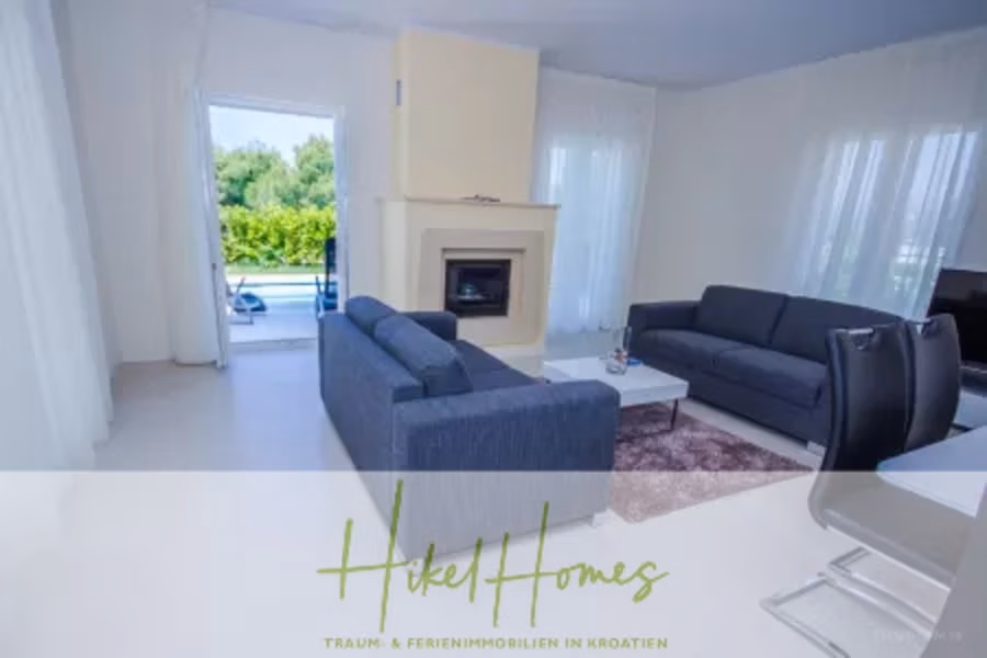 Villa im Mittelmeerstil mit 3 SZ, Pool, Dachterrasse mit Meerblick (2) - Ein modernes Wohnzimmer mit zwei dunklen Sofas vor einem Kamin. Ein Couchtisch und ein Teppich stehen dazwischen. Große Fenster lassen Tageslicht herein, und draußen bietet sich ein Blick auf Pool und Grün. Das Bild trägt den Text „Hikel Homes“. (41881)