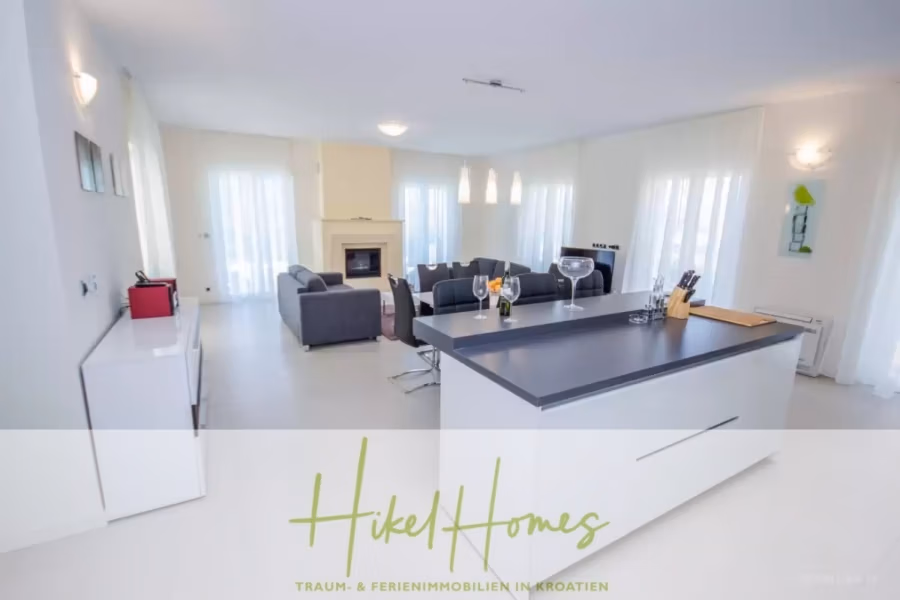 Villa im Mittelmeerstil mit 3 SZ, Pool, Dachterrasse mit Meerblick (2) - Modernes, helles Wohnzimmer und Küche mit weißen Wänden und großen Fenstern. Die Kücheninsel verfügt über eine schwarze Arbeitsplatte mit Weingläsern und einem Schneidebrett. Eine graue Sofagarnitur steht neben einem Kamin. Von der Decke hängen Lampen. (41880)