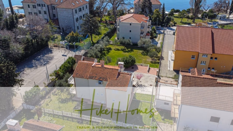 300m² Villa in 2. Reihe zum Meer mit großem Grundstück mit Erweiterungspotential - Luftaufnahme eines Wohngebiets nahe der Küste mit mehreren Häusern mit roten Ziegeldächern. Im Hintergrund ist das Meer zu sehen. Der Textüberlagerungstext lautet „Hikel Homes“ mit zusätzlichem kleineren Text darunter. (41842)