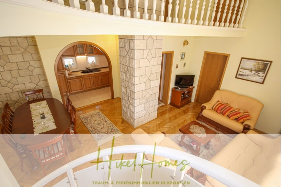 300m² Villa in 2. Reihe zum Meer mit großem Grundstück mit Erweiterungspotential - Gemütlicher Wohnbereich mit beigem Sofa und Sesseln, einem kleinen Fernseher auf einem Holzständer und einem Esstisch mit dunklen Stühlen. Ein Torbogen führt in die Küche mit Holzschränken. Der Raum ist mit Stein- und Holzelementen gestaltet und verfügt über eine Treppe und ein Balkongeländer darüber. (41849)