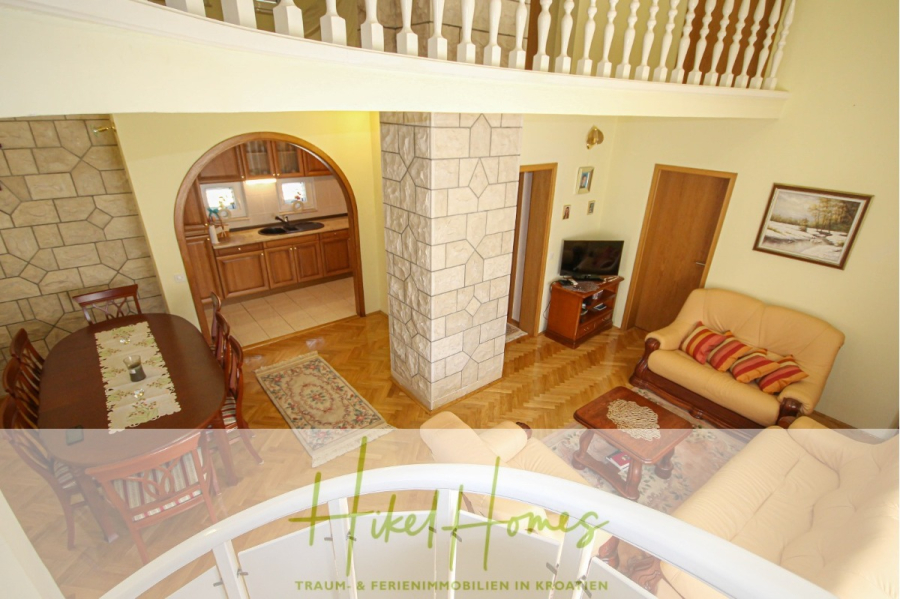 300m² Villa in 2. Reihe zum Meer mit großem Grundstück mit Erweiterungspotential - Geräumiger Wohn- und Essbereich mit beigen Sofas, Holzmöbeln, TV und einer zentralen Säule. Ein gewölbter Durchgang führt in die Küche. Das Balkongeländer im Obergeschoss ist sichtbar. Das Logo "Hikel Homes" erscheint am unteren Rand. (44960)