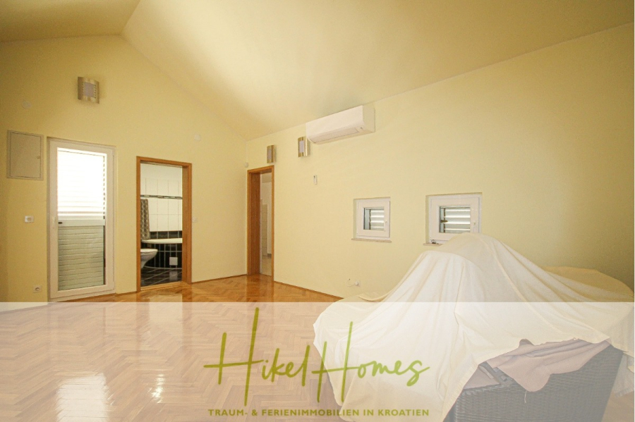 300m² Villa in 2. Reihe zum Meer mit großem Grundstück mit Erweiterungspotential - Ein helles, geräumiges Zimmer mit hellgelben Wänden, Holzparkett, einer Klimaanlage, zwei kleinen Fenstern und einem unbedeckten Eingang zum Bad. Die Möbel sind mit weißen Tüchern bedeckt. Logo "Hikel Homes" am Boden. (44971)