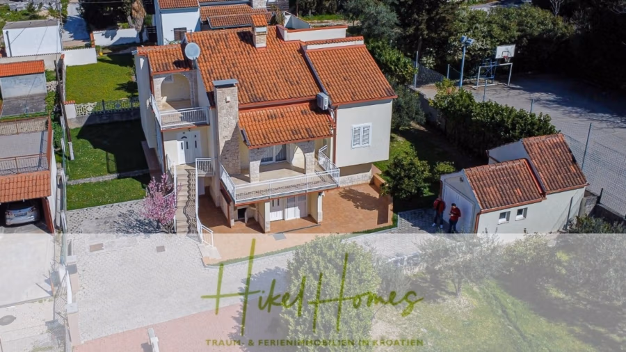 300m² Villa in 2. Reihe zum Meer mit großem Grundstück mit Erweiterungspotential - Luftaufnahme eines zweistöckigen Hauses mit rotem Ziegeldach, umgeben von Grünflächen und einer Auffahrt. In der Nähe befinden sich ein Basketballkorb und ein kleines Nebengebäude. Im Vordergrund befindet sich eine halbtransparente Überlagerung mit dem Text „Hikel Homes“. (41841)