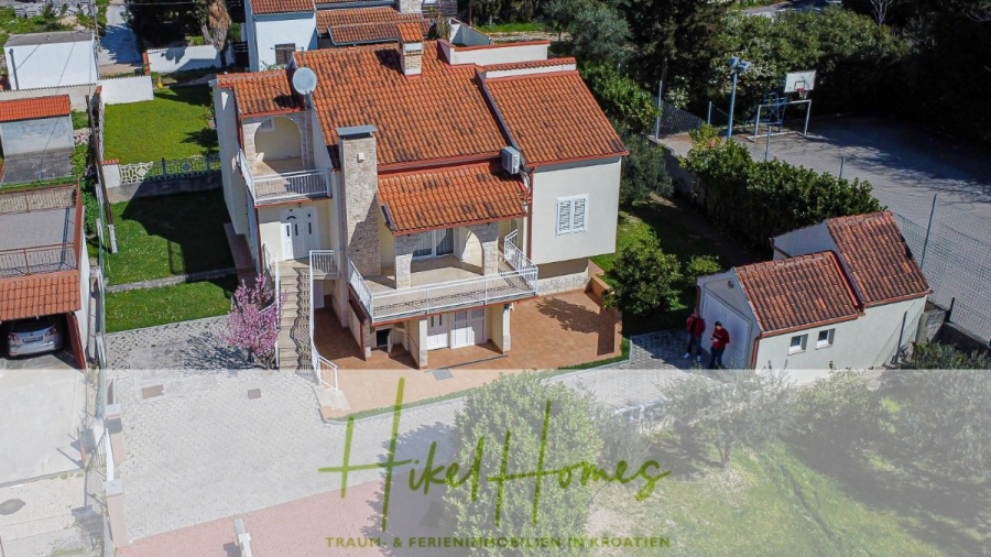 300m² Villa in 2. Reihe zum Meer mit großem Grundstück mit Erweiterungspotential - Luftaufnahme eines zweistöckigen Hauses mit roten Ziegeldächern, weißen Wänden, Balkonen, einem Schornstein und einem kleinen freistehenden Gebäude, umgeben von Grünflächen und einer gepflasterten Auffahrt. Der überlagerte Text lautet "Hikell Homes". (44951)