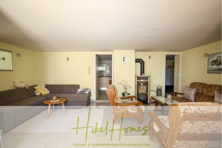 300m² Villa in 2. Reihe zum Meer mit großem Grundstück mit Erweiterungspotential - Gemütliches Wohnzimmer mit zwei Sofas, Sesseln, einem Holzofen, Couchtisch und Teddybären. Neutrale Wände, ein offener Durchgang zur Küche und Kunstwerke schmücken den Raum. Der Text am Boden lautet: "Hikel Homes Traum- & Ferienimmobilien in Kroatien. (44963)
