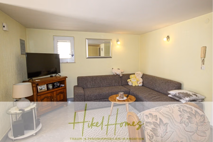 300m² Villa in 2. Reihe zum Meer mit großem Grundstück mit Erweiterungspotential - Gemütliches Wohnzimmer mit einem grauen L-förmigen Sofa, geschmückt mit Teddybären, einem hölzernen TV-Schrank mit Flachbildfernseher, einem runden Beistelltisch aus Holz und einem Wandspiegel. Sanfte Beleuchtung und grüne Wände schaffen eine warme Atmosphäre. Unten ist das Logo „Hikel Homes“ zu sehen. (41844)