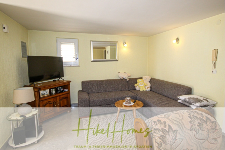 300m² Villa in 2. Reihe zum Meer mit großem Grundstück mit Erweiterungspotential - Ein gemütliches Wohnzimmer mit einem grauen L-förmigen Sofa, Teddybären, einem Flachbildfernseher auf einem Holzständer, einem runden Beistelltisch aus Glas und blassgelben Wänden mit Wandleuchten und einem Spiegel. (44954)