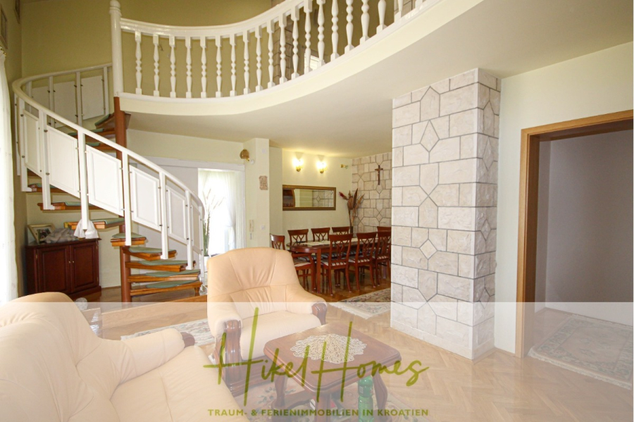 300m² Villa in 2. Reihe zum Meer mit großem Grundstück mit Erweiterungspotential - Geräumiger Wohn- und Essbereich mit beigen Sesseln, Esstisch aus Holz, Akzentwand aus Stein und einer weißen Wendeltreppe, die zu einem offenen Balkon führt. Große Fenster sorgen für natürliches Licht. (44959)