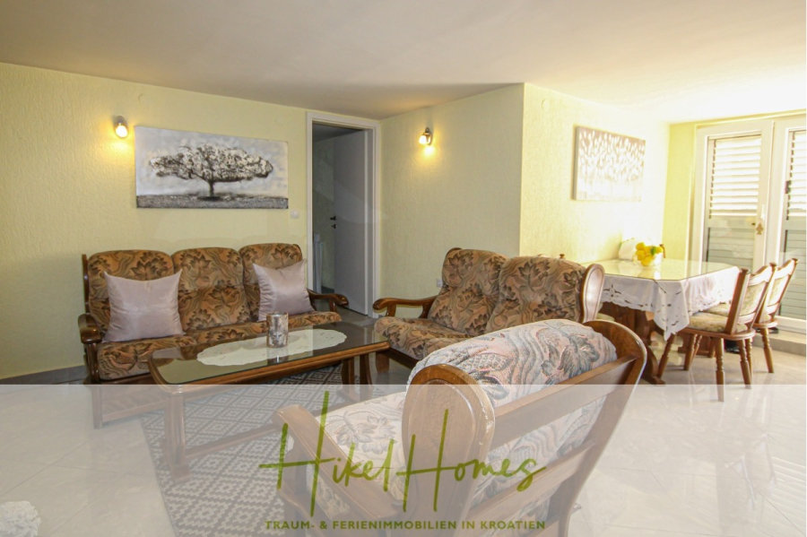 300m² Villa in 2. Reihe zum Meer mit großem Grundstück mit Erweiterungspotential - Ein gemütliches Wohnzimmer mit blumengemusterten Sofas, einem Couchtisch aus Glas, einem an der Wand befestigten Baumkunstwerk und einem Essbereich mit Tisch und Stühlen in der Nähe der Fensterläden. Das Logo von "Hikel Homes" ist am unteren Rand eingeblendet. (44962)