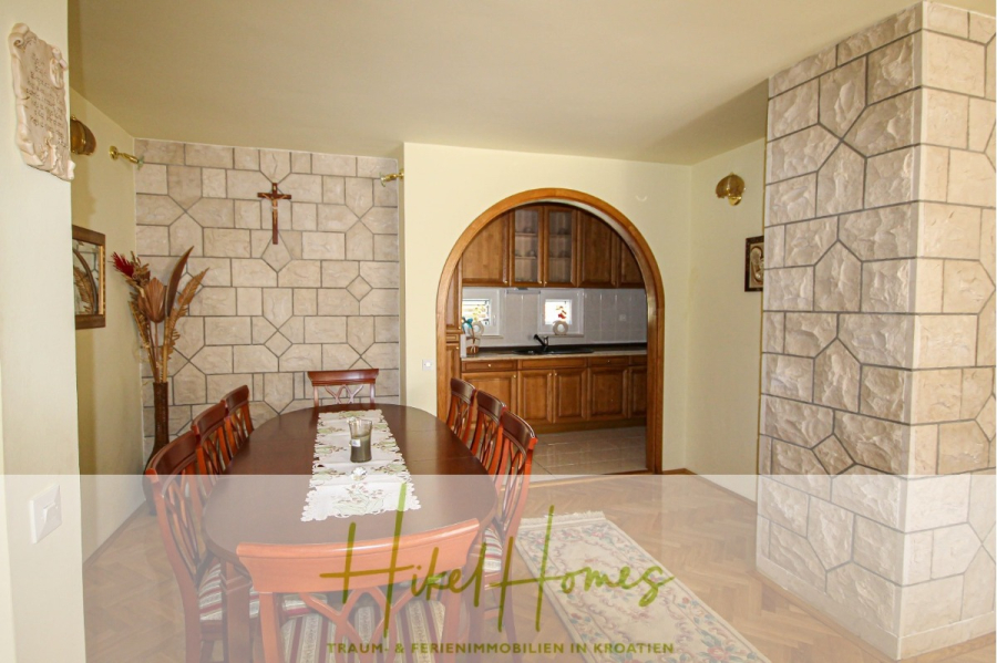 300m² Villa in 2. Reihe zum Meer mit großem Grundstück mit Erweiterungspotential - Ein Esszimmer mit einem langen Holztisch und Stühlen, mit Steinmustern verzierten Wänden und einer gewölbten Tür, die zu einer Küche mit Holzschränken führt. Der Text "Hike Homes" ist am unteren Rand eingeblendet. (44958)