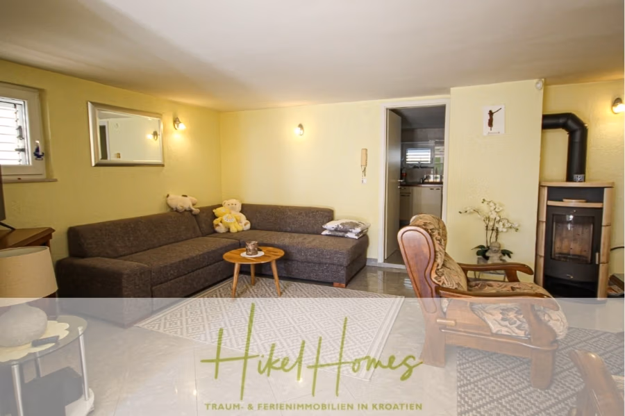 300m² Villa in 2. Reihe zum Meer mit großem Grundstück mit Erweiterungspotential - Gemütliches Wohnzimmer mit einem grauen Sofa, einem Couchtisch aus Holz und einem Vintage-Sessel. In der Ecke befindet sich ein kleiner Kamin. Über dem Sofa hängt ein Spiegel, im Hintergrund ist eine kleine Küchenzeile zu sehen. Der Raum ist sanft und warm beleuchtet. (41858)