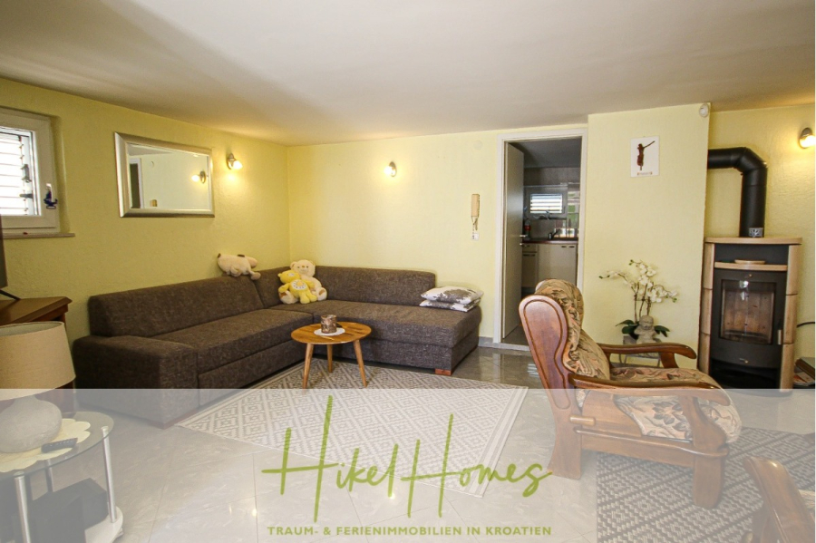 300m² Villa in 2. Reihe zum Meer mit großem Grundstück mit Erweiterungspotential - Ein gemütliches Wohnzimmer mit einem grauen Sektionssofa, einem runden hölzernen Couchtisch, einem gepolsterten Sessel und einem holzbefeuerten Ofen. Eine kleine Küche ist durch eine offene Tür sichtbar. Der überlagerte Text lautet: "Hikel Homes. (44972)