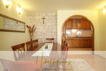 Ein Esszimmer mit einem langen Holztisch und sechs Stühlen, einem dekorativen Wandspiegel und einem Pflanzenarrangement. Im Hintergrund führt eine gewölbte Tür zu einer Küche mit Holzschränken. - 300m² Villa in 2. Reihe zum Meer mit großem Grundstück mit Erweiterungspotential