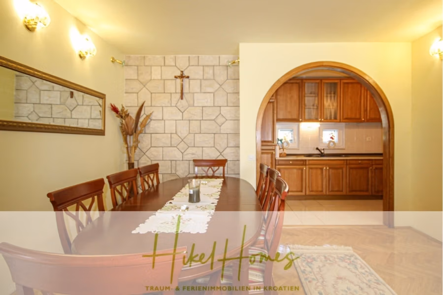 300m² Villa in 2. Reihe zum Meer mit großem Grundstück mit Erweiterungspotential - Esszimmer mit langem Holztisch und Stühlen. Ein dekoratives Tuch ist über den Tisch gespannt. Die Wände sind hell gestrichen und mit Steinakzenten versehen. Ein Torbogen führt in die Küche mit Holzschränken. Ein Kreuz und Pflanzen schmücken die Wände. Unten ist der Text „HikeHomes“ eingelegt. (41845)