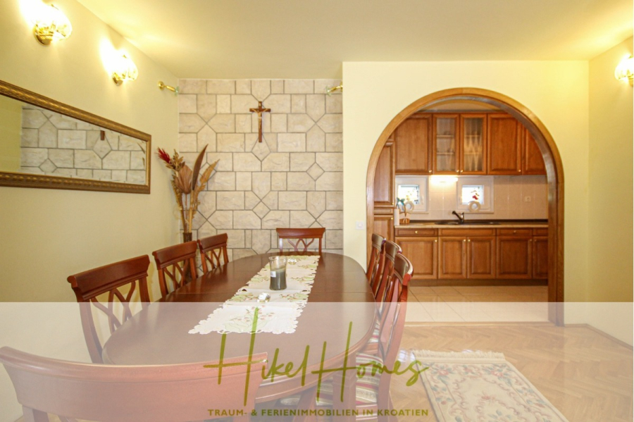 300m² Villa in 2. Reihe zum Meer mit großem Grundstück mit Erweiterungspotential - Ein Esszimmer mit einem langen Holztisch und sechs Stühlen, einem dekorativen Wandspiegel und einem Pflanzenarrangement. Im Hintergrund führt eine gewölbte Tür zu einer Küche mit Holzschränken. (44955)