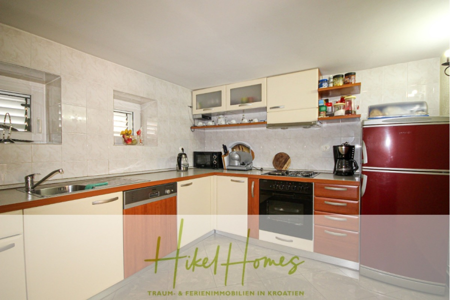 300m² Villa in 2. Reihe zum Meer mit großem Grundstück mit Erweiterungspotential - Moderne Küche mit cremefarbenen und hölzernen Schränken, eingebautem Ofen, Herd, rotem Kühlschrank, Spüle und verschiedenen Geräten auf der Arbeitsplatte. Über der Spüle befindet sich ein Fenster, und die Wände sind gefliest. Der Text des Logos ist unten überlagert. (44964)