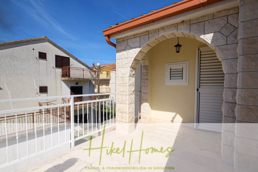 300m² Villa in 2. Reihe zum Meer mit großem Grundstück mit Erweiterungspotential - Ein sonnendurchflutetes Steinhaus mit Ziegeldach und gewölbtem Eingang mit Laterne und geschlossenen Fensterläden. Ein weißes Metallgeländer blickt auf die Nachbarhäuser. Der Text lautet „Hikel Homes – Traum- & Ferienimmobilien in Kroatien“. (41860)