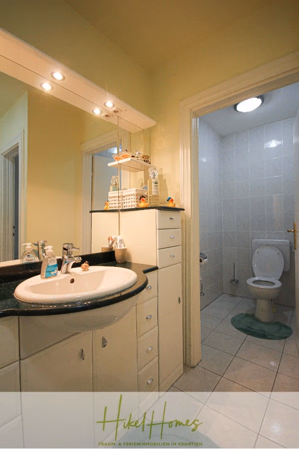 300m² Villa in 2. Reihe zum Meer mit großem Grundstück mit Erweiterungspotential - Ein kleines Badezimmer mit Waschbecken und Ablagefläche auf der linken Seite, Toilettenartikeln und einem beleuchteten Spiegel. Eine Tür führt zum Toilettenbereich mit weiß gefliesten Wänden und Boden, einer grünen Matte und einem Regal mit Dekorationen. (41856)