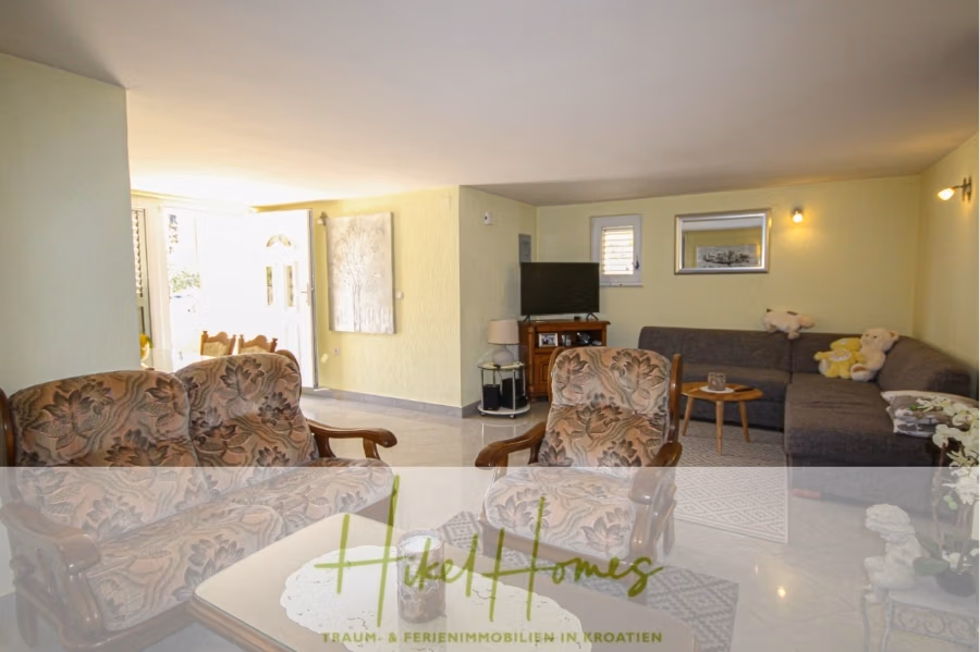 300m² Villa in 2. Reihe zum Meer mit großem Grundstück mit Erweiterungspotential - Ein gemütliches Wohnzimmer mit floral gemusterten Sesseln, einem großen grauen Sofa, einem Flachbildfernseher und dekorativen Kissen. Der Raum verfügt über helle Wände, einen Essbereich und sanfte Beleuchtung. Unten ist das Logo „Hikel Homes“ zu sehen. (41846)