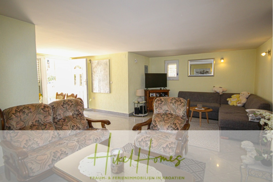 300m² Villa in 2. Reihe zum Meer mit großem Grundstück mit Erweiterungspotential - Helles Wohnzimmer mit gemusterten Sesseln, einem grauen Sofa, einem Fernseher auf einem Ständer und großen Fenstern, die natürliches Licht hereinlassen. Auf dem Sofa befinden sich Dekorationsartikel und Stofftiere. Das Logo "Hike Homes" erscheint am unteren Rand. (44956)