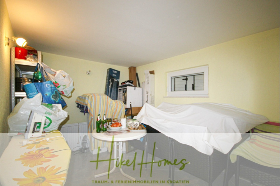 300m² Villa in 2. Reihe zum Meer mit großem Grundstück mit Erweiterungspotential - Ein unordentlicher Raum mit abgedeckten Möbeln, Taschen, Flaschen und Haushaltsgegenständen, die auf Regalen und Tischen gestapelt sind. An der rechten Wand befindet sich ein kleines Fenster, und der Raum ist hell erleuchtet mit gelben Wänden. Der Schriftzug "Hikel Homes" überlagert das Bild. (44966)