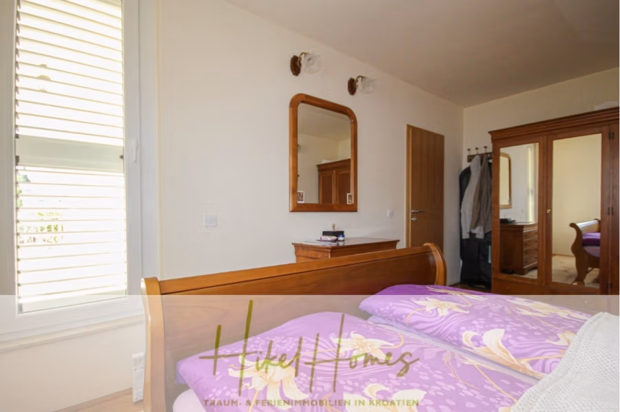 300m² Villa in 2. Reihe zum Meer mit großem Grundstück mit Erweiterungspotential - Schlafzimmer mit einem Holzbett mit lila geblümter Bettwäsche. Über einer kleinen Kommode hängt ein Spiegel an der Wand. Rechts befinden sich ein Kleiderschrank und hängende Kleidung. Links ein Fenster mit weißen Jalousien. Unten steht „Hicke Homes“. (41862)