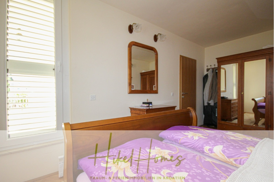 300m² Villa in 2. Reihe zum Meer mit großem Grundstück mit Erweiterungspotential - Ein helles Schlafzimmer mit einem Holzbett und lila geblümter Bettwäsche, einer kleinen Kommode mit Spiegel, einem Holzkleiderschrank und einem Fenster mit weißen Fensterläden. Das Logo "Hohe Homes" erscheint am unteren Rand. (44978)