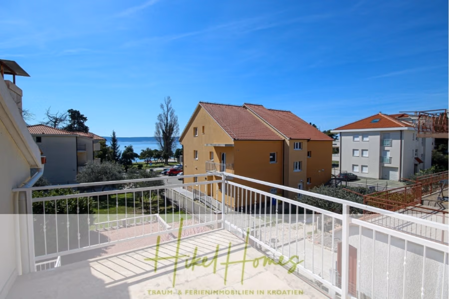 300m² Villa in 2. Reihe zum Meer mit großem Grundstück mit Erweiterungspotential - Blick vom Balkon auf einen sonnigen Tag mit klarem, blauem Himmel. Beige und gelbe Häuser, Bäume und ein Blick auf das Meer im Hintergrund. Ein weißes Geländer säumt den Balkon. Die Textüberlagerung lautet „Exklusive Homes – Traum-Ferienimmobilien in Kroatien“. (41859)