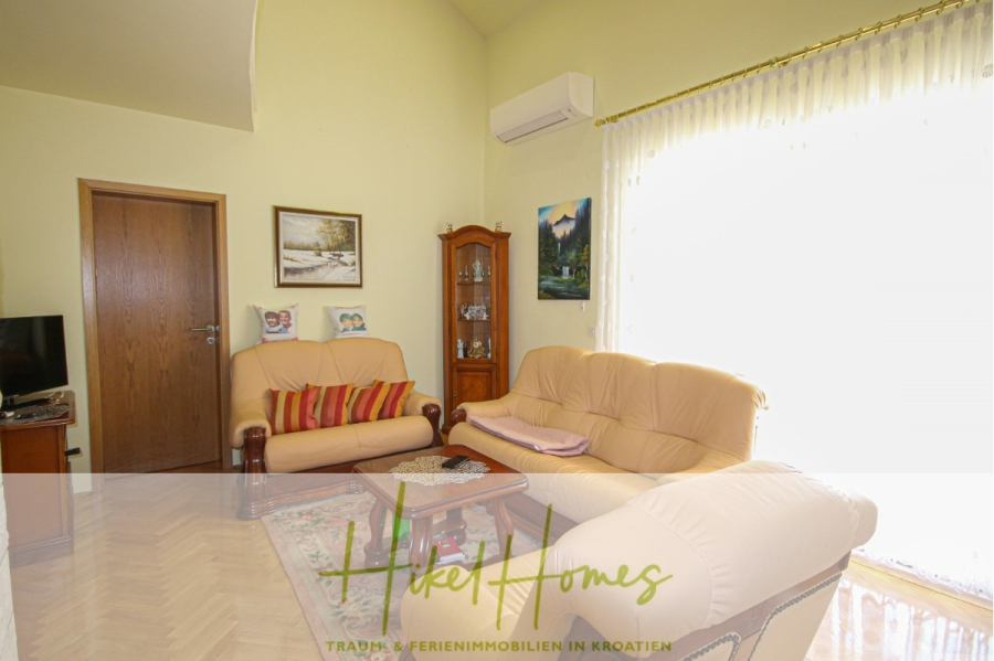 300m² Villa in 2. Reihe zum Meer mit großem Grundstück mit Erweiterungspotential - Ein gemütliches Wohnzimmer mit cremefarbenen Sofas, einem hölzernen Couchtisch, einer Glasvitrine, Wandkunst und großen Fenstern mit transparenten Vorhängen, die natürliches Licht hereinlassen. (44968)