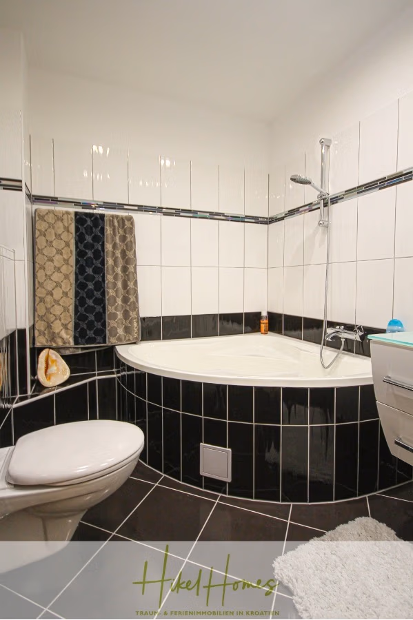300m² Villa in 2. Reihe zum Meer mit großem Grundstück mit Erweiterungspotential - Modernes Badezimmer mit Eckbadewanne und Dusche, weiß-schwarz gefliesten Wänden und dunklen Bodenfliesen. Handtücher hängen an einem Ständer über der Wanne. Links befindet sich eine Toilette mit einer Badematte auf dem Boden. (41854)