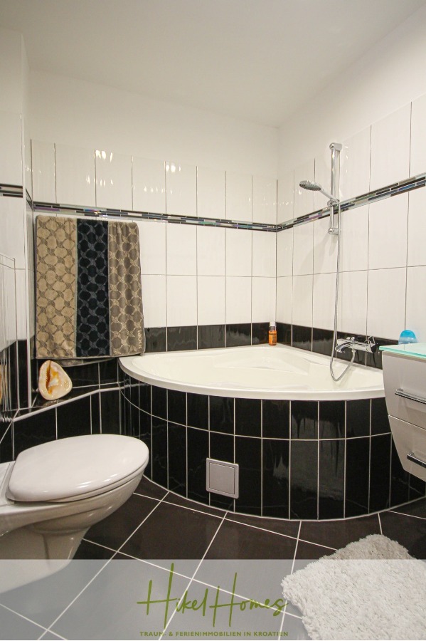 300m² Villa in 2. Reihe zum Meer mit großem Grundstück mit Erweiterungspotential - Modernes Badezimmer mit schwarzen und weißen Fliesen, einer Eckbadewanne mit Dusche, einer Toilette, einem Handtuchhalter mit zwei Handtüchern und einem kleinen Ablagefach. Auf dem Boden liegt eine graue Badematte. (44967)