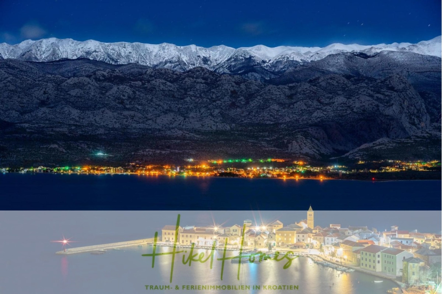 Luxus-Meerblick und Gebirgsblick-Villa in 2. Reihe zum Meer mit 4 SZ, Pool, Sauna, Fitness & Jacuzzi - Eine kroatische Küstenstadt bei Nacht mit beleuchteten Gebäuden entlang der Uferpromenade. Im Hintergrund erheben sich schneebedeckte Berge unter einem klaren, sternenklaren Himmel. Der Text am unteren Rand lautet "Hike Homes - Traum- & Ferienimmobilien in Kroatien. (46039)