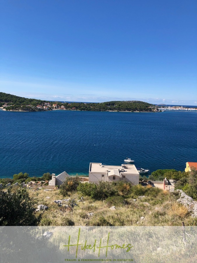 Einmalige 2. Reihe mit Meerzugang – 1834m² Bauland mit Baugenehmigung für 2 Villen - Blick auf ein tiefblaues Meer mit einigen Booten in Ufernähe, Häusern und Grünflächen im Vordergrund und einer baumbewachsenen Insel in der Ferne unter einem klaren blauen Himmel. (44061)