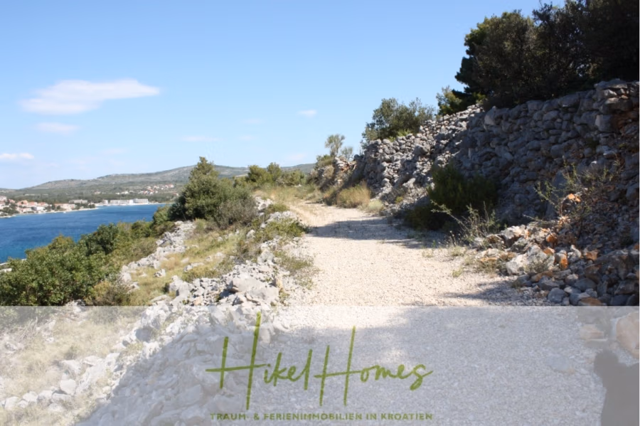 Einmaliges 2. Reihe mit Meerzugang 1834m² Bauland mit Baugenehmigung für 2 Villen - Ein schmaler Schotterweg schlängelt sich an einem felsigen Hang entlang, mit Blick auf das Meer zur Linken. Spärliche Vegetation und Bäume säumen den Weg unter einem strahlend blauen Himmel. Am Ende befindet sich ein Wasserzeichen mit der Aufschrift „Hikel Homes“. (43218)