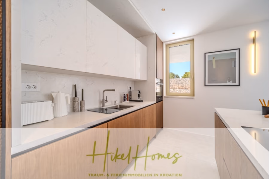 Hochwertige Meerblick Villa SUN in Sutivan (Insel Brac) – 3 SZ + Bad, Pool, Sommerküche, WC, … - Moderne Küche mit eleganten weißen Schränken und Arbeitsplatten. Zur Ausstattung gehören eine Spüle, ein Toaster und eine Espressomaschine. Ein Fenster sorgt für Tageslicht. Ein gerahmtes Kunstwerk und eine vertikale Leuchte schmücken die Wände. Der Boden ist hell. (42966)