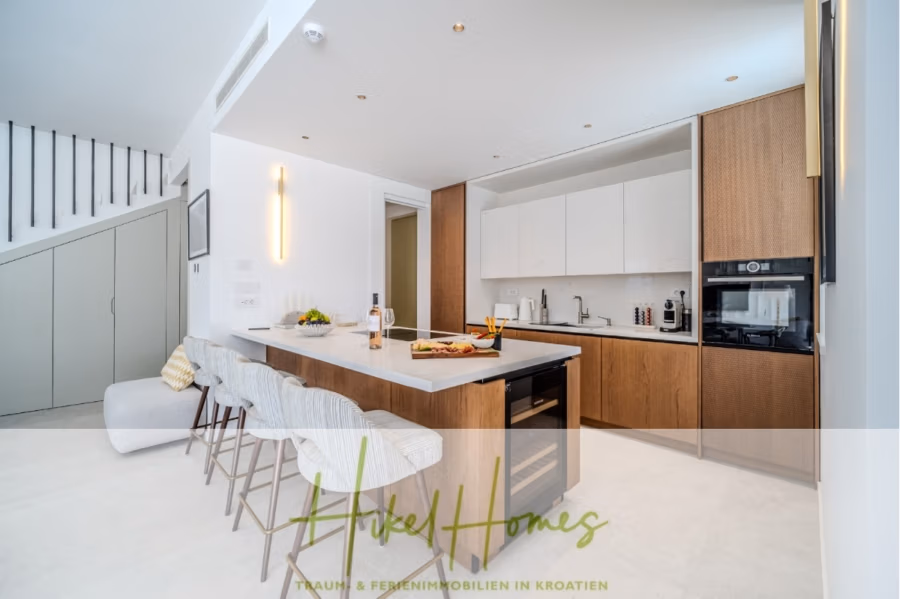 Hochwertige Meerblick Villa SUN in Sutivan (Insel Brac) – 3 SZ + Bad, Pool, Sommerküche, WC, … - Moderne Küche in hellem Farbton mit Marmor-Arbeitsinsel mit Hockern, braunen Schränken, Einbaubackofen und Spüle. Im Hintergrund sind Treppe und Stauraum zu sehen. Zur Dekoration gehören Flaschen und Obst auf der Kücheninsel. (42968)